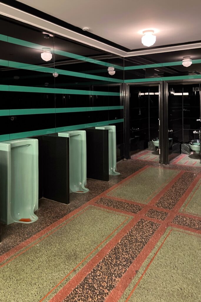 Men’s restroom at Drusie & Darr inside the Hermitage Hotel in Nashville, with vintage mint-green urinals, glossy black and emerald striped tile walls, terrazzo flooring in green and rust tones, and retro light fixtures — a preserved example of historic hotel bathroom design.