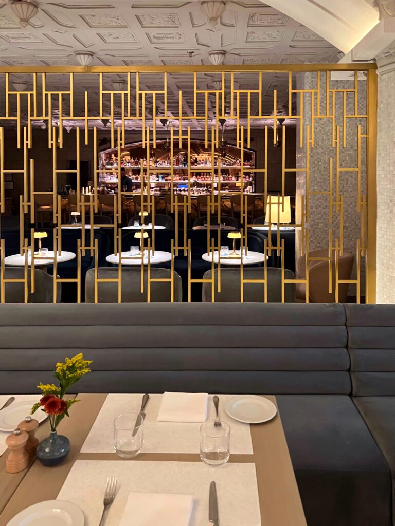 Dining area at Drusie & Darr inside the Hermitage Hotel in Nashville, featuring a contemporary brass metal screen partition, upholstered banquette seating, and a view through to the marble bar with illuminated shelves beneath a coffered ceiling — a layered example of restaurant interior design.