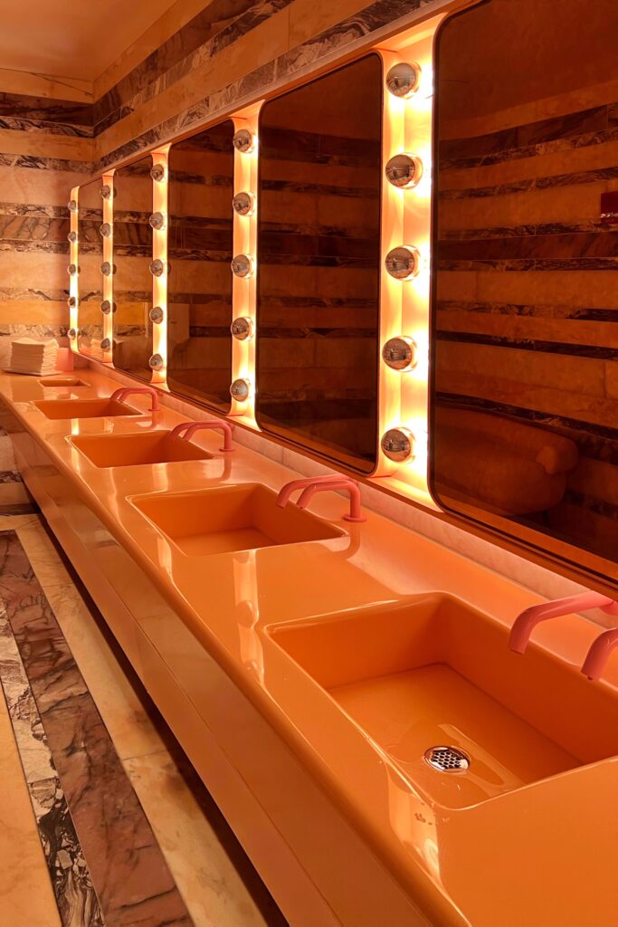 Women’s bathroom at Drusie & Darr inside the Hermitage Hotel in Nashville, featuring glossy pink sinks vanity with pink faucets, a dramatic backlit mirror framed with globe bulbs, and striped marble walls for a retro-modern interior design statement.
