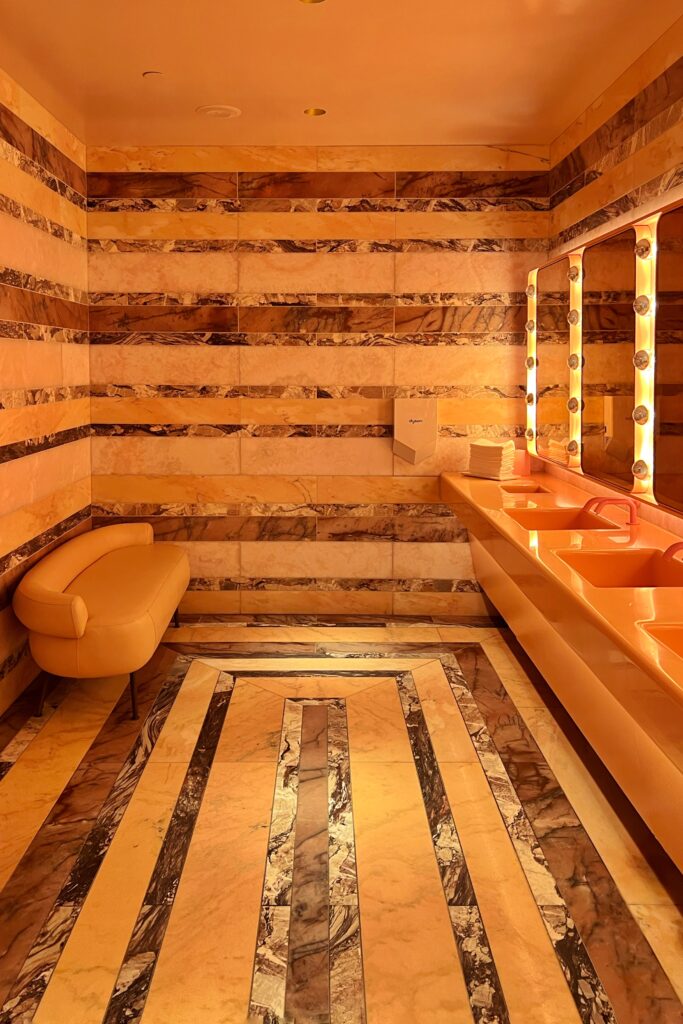 Women’s bathroom at Drusie & Darr in the Hermitage Hotel, Nashville, showcasing striped marble walls and geometric marble flooring, pink vanities with pink faucets, illuminated mirrors, and a curved upholstered bench — an iconic example of Nashville interior design.