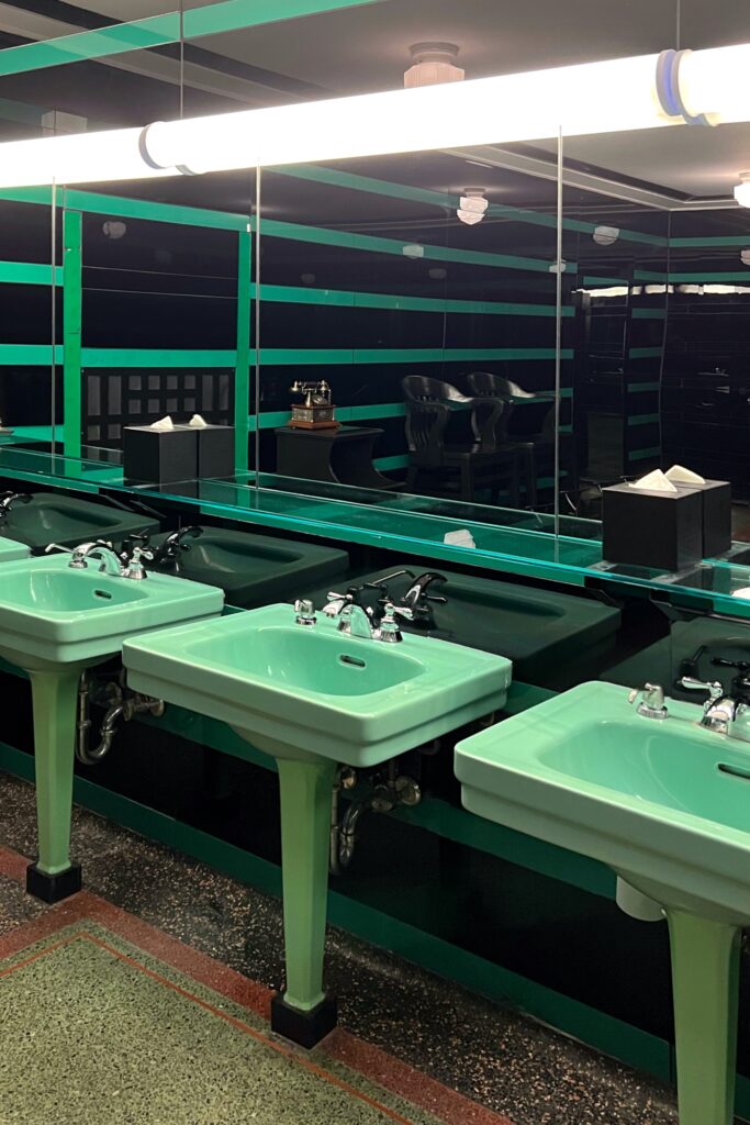 Men’s restroom at Drusie & Darr inside the Hermitage Hotel in Nashville, featuring vintage mint-green pedestal sinks, glossy black and green striped walls, terrazzo flooring, and retro light fixtures — an iconic example of historic hotel bathroom interior design.”