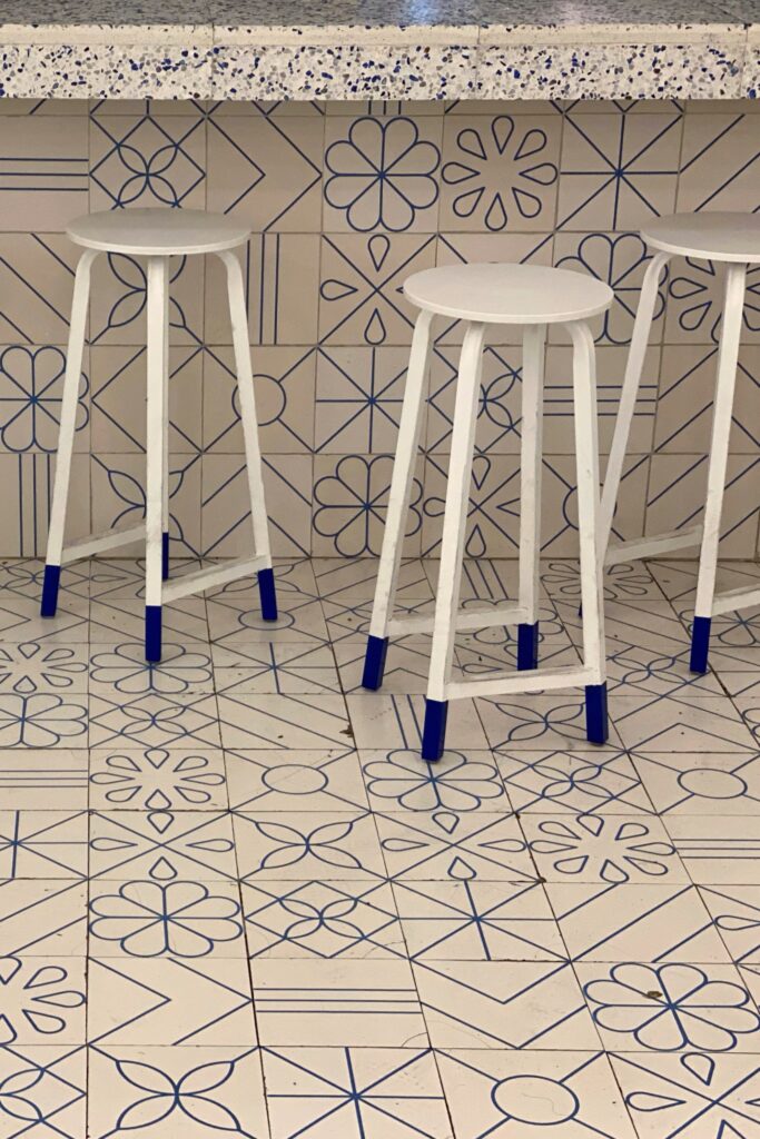 White bar stools with blue-dipped legs set against patterned blue-and-white tiled walls, a blue-and-white terrazzo bar top, and graphic tile floors at El Moro Churrería in Mexico City.