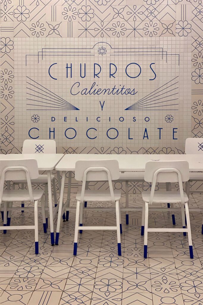 White tiled wall with blue lettering at El Moro Churrería in Mexico City, reading 'Churros Calientitos y Delicioso Chocolate,' with white tables and chairs in the seating area.