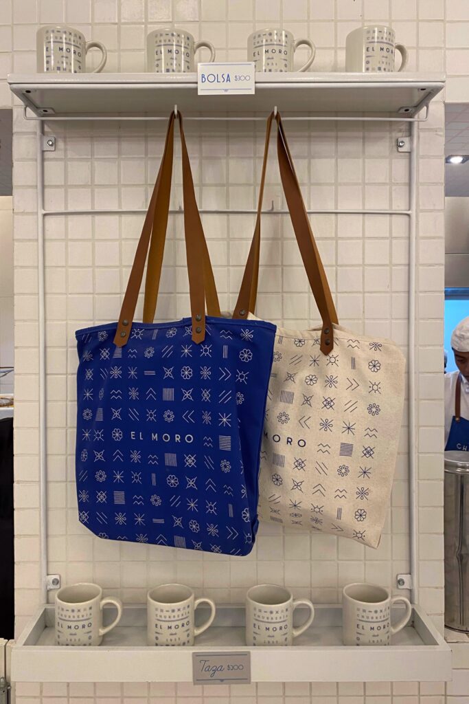 Tote bags and mugs from El Moro, the iconic churrería in Mexico City, displayed against a white tiled wall with bold geometric blue designs and leather strap details.