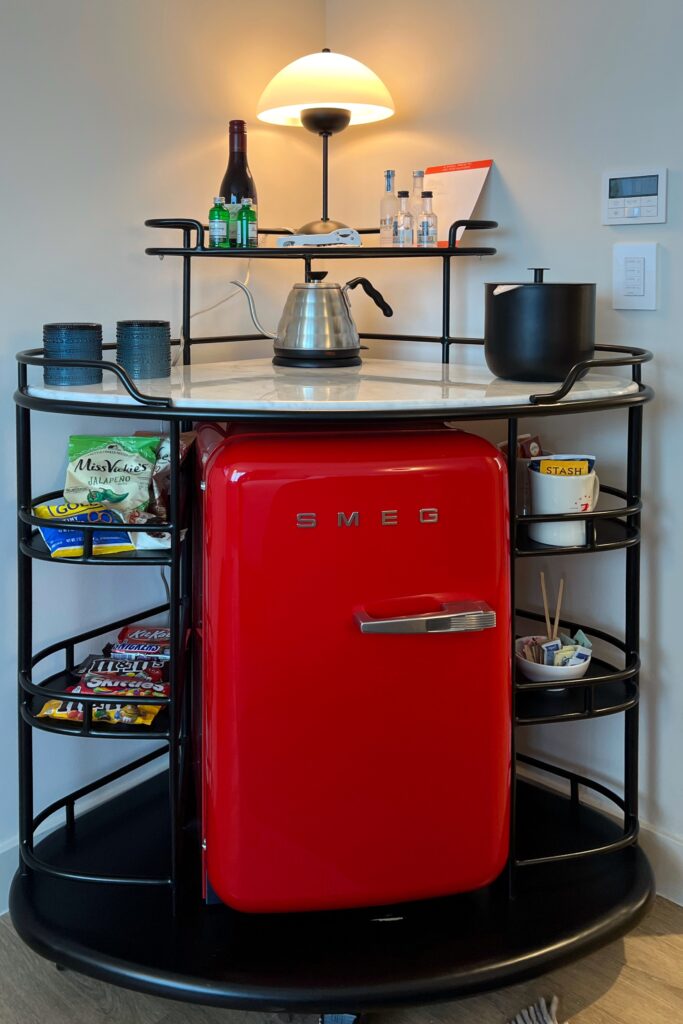 Stylish hotel minibar at Virgin Hotel Nashville featuring a retro red Smeg fridge, marble top, modern black frame, and curated minibar snacks.