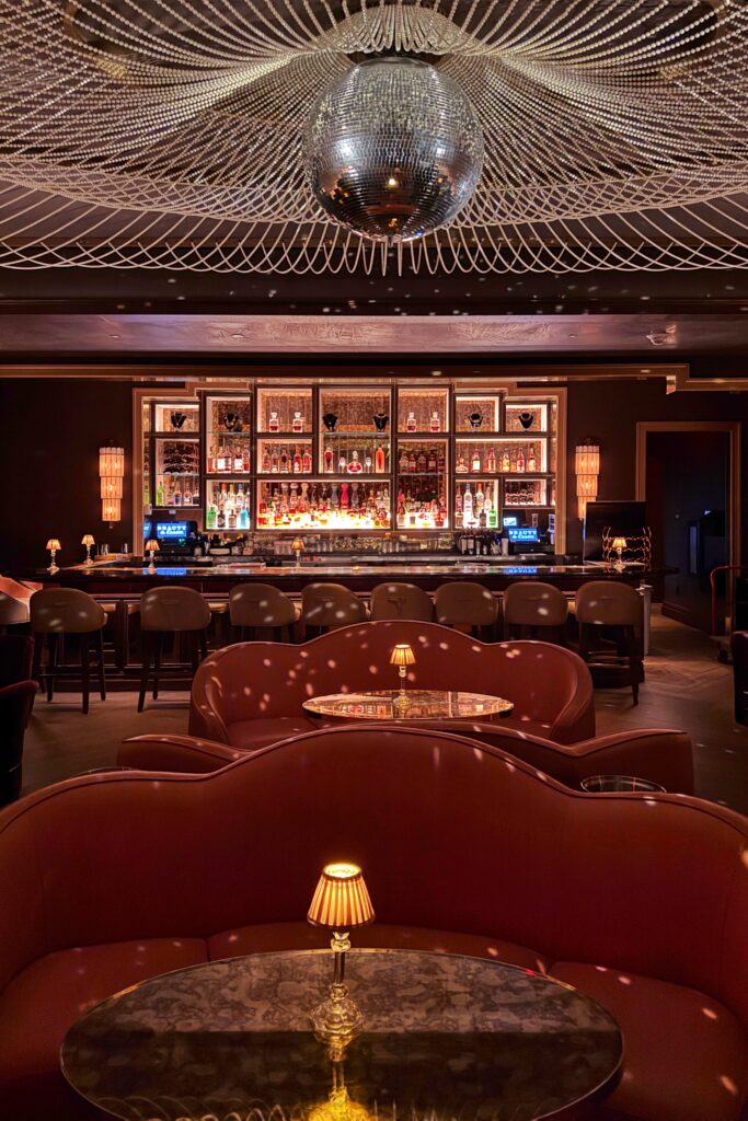 Beauty & Essex speakeasy bar restaurant interior featuring scalloped leather booth seating, marble-topped tables with miniature pleated lamps, an illuminated back bar of glass bottles, and a dramatic disco ball suspended beneath a beaded ceiling.
