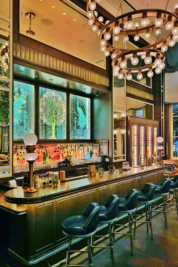 Modern Art Deco bar interior at Café Carmellini in New York City, featuring teal glass peacock panels, brass accents, globe chandeliers, and blue leather stools that evoke modern luxury and speakeasy style elegance.