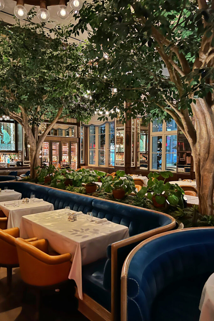 Elegant modern Art Deco dining room at Café Carmellini in New York City, featuring lush indoor trees, long linear blue velvet banquettes, and brass accents that create a luxurious modern speakeasy style atmosphere.