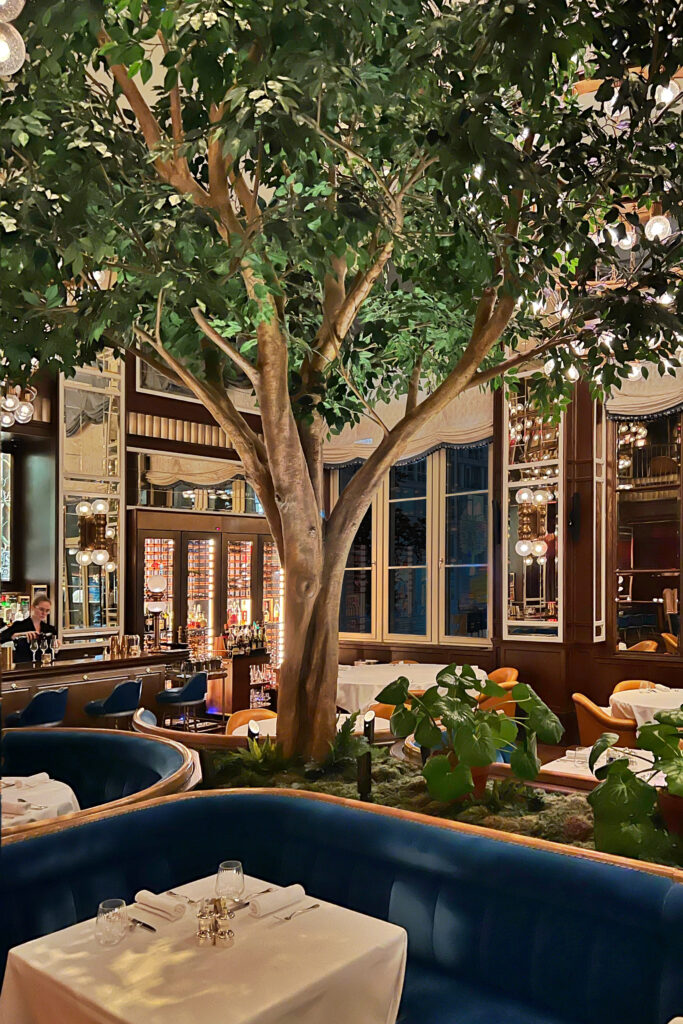 The lush, modern Art Deco interior of Café Carmellini in New York City, featuring a dramatic indoor tree, mirrored walls, velvet banquettes, and brass lighting that create an elegant fine dining atmosphere.