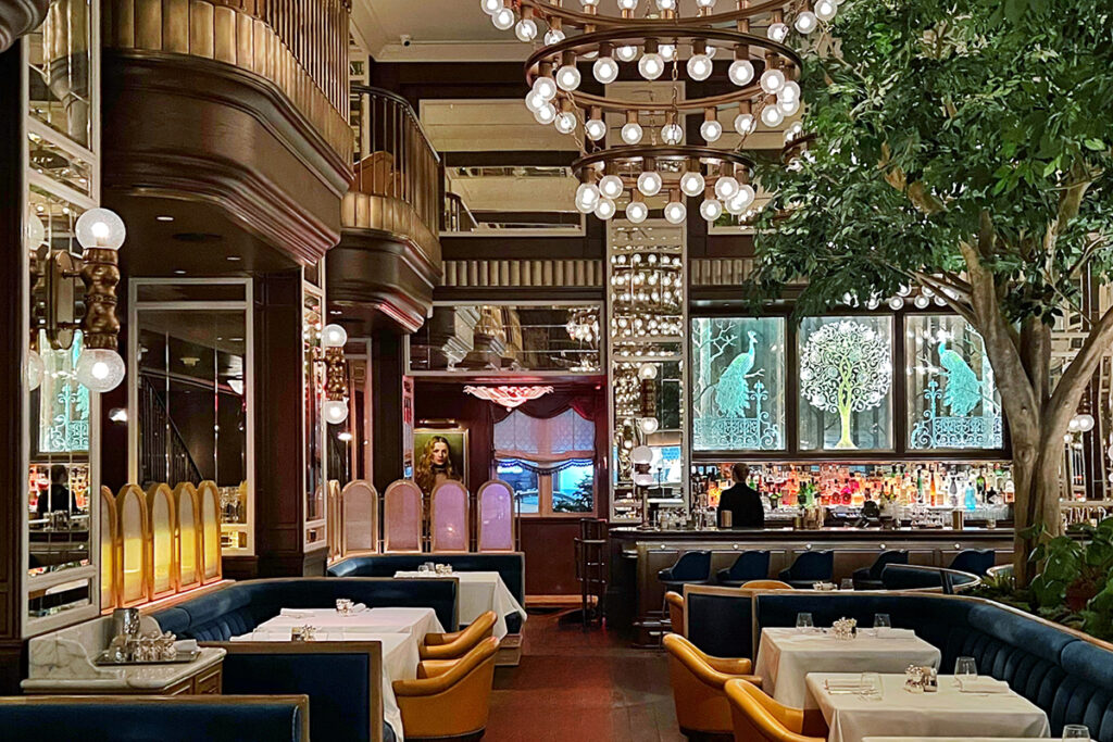 Opulent interior of Café Carmellini in New York City featuring tiered circular chandeliers with glowing globe bulbs, dark wood paneling, brass balcony details, deep blue tufted banquettes, mustard leather armchairs, white tablecloths, a mirrored back bar, and illuminated peacock and tree artwork—an aesthetic NYC restaurant that blends grand European brasserie style with theatrical design for food lovers and design-focused travelers.