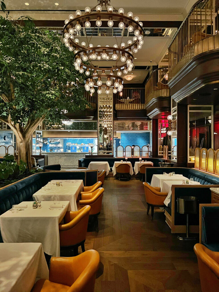 Elegant Modern Art Deco dining room at Café Carmellini restaurant in New York City, featuring circular chandeliers, brass balconies, rich blue velvet banquettes, and golden leather chairs that create a luxurious modern restaurant interior.