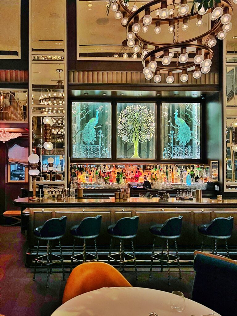 The elegant Modern Art Deco bar at Café Carmellini in New York City, featuring illuminated peacock glass panels, brass lighting, and jewel-toned velvet barstools that highlight the restaurant’s modern luxury design.