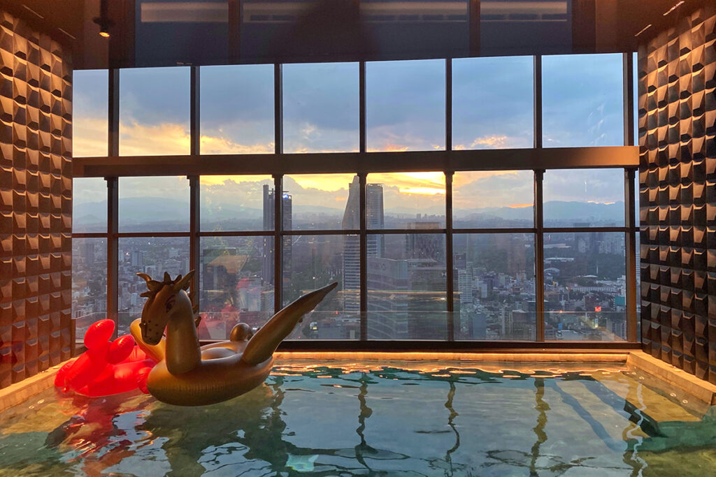 Rooftop pool at Sofitel Mexico City Reforma during sunset, with sculptural wall textures, inflatable pool floaties, and panoramic skyline views through floor-to-ceiling windows, creating a modern and luxurious city escape.