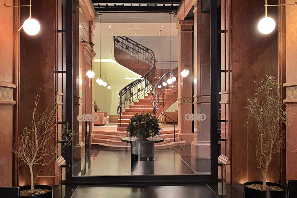 Entrance view of Hotel Umbral in Mexico City featuring a grand curved staircase with globe pendant lights, warm stone, and modern glass doors framed by architectural columns, blending historic detail with contemporary design.