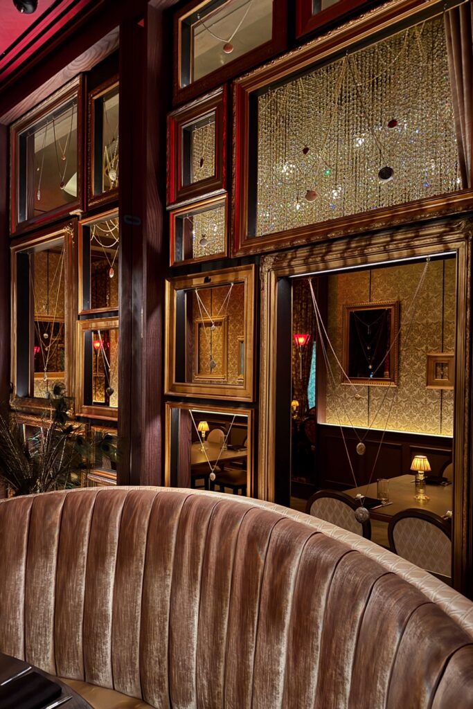 Beauty & Essex interior featuring gold-framed mirrors and hanging jewelry necklaces, velvet banquette seating, and moody Art Deco lighting inspired by speakeasy design.