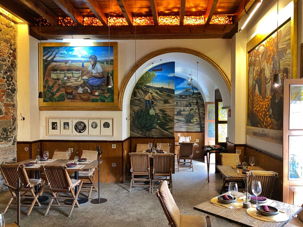 The upstairs dining area at Limosneros restaurant in Mexico City features hand-painted murals, arched doorways, and rustic wood furniture under a beamed ceiling — a warm, artful interior featured in the Roam & Reside Mexico City Aesthetic Travel Guide.