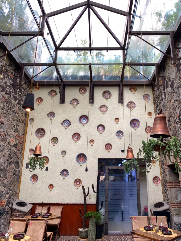 Atrium dining space at Los Limosneros restaurant in Mexico City’s Centro Histórico, featuring a glass ceiling, volcanic stone walls, and a white accent wall decorated with colorful Talavera plates. Hanging copper lights, potted cacti, and woven chairs complete the look — a bright, design-forward restaurant interior that captures modern Mexican style and craftsmanship in Mexico City.