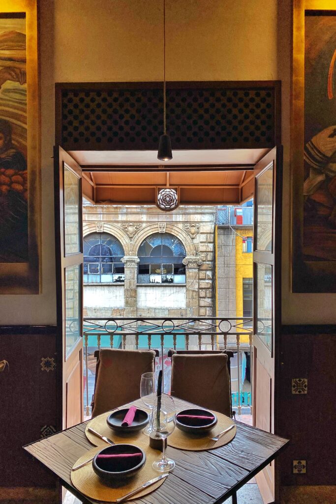 Balcony dining table at Los Limosneros restaurant in Mexico City’s Centro Histórico, overlooking the ornate stone arches of a historic building across the street. The intimate setting features a rustic wooden table set with minimalist Mexican dishware and gold placemats, blending fine dining with old-world architecture — an aesthetic Mexico City restaurant experience.