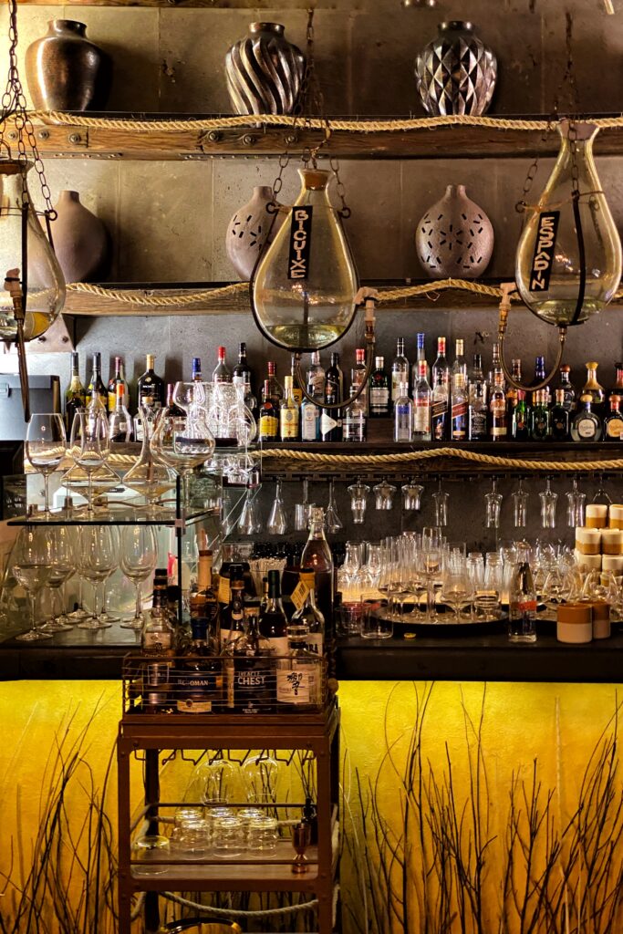 Warm, moody bar interior at Los Limosneros in Mexico City’s Centro Histórico, featuring artisanal mezcal bottles, glass decanters suspended with rope, and hand-thrown ceramic vases on rustic wooden shelves. A design-forward cocktail bar blending contemporary Mexican craftsmanship, ambient lighting, and industrial-chic decor — ideal for travelers seeking the best bars and restaurants in Mexico City.