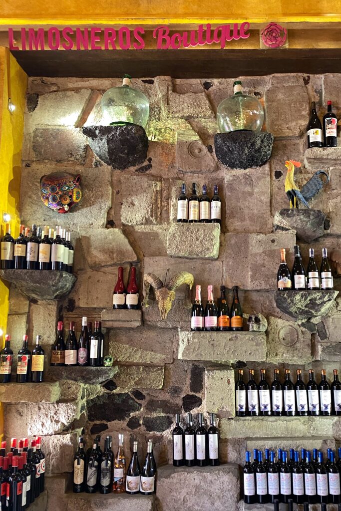 Stone wall wine display at Los Limosneros Boutique with pink signage in Mexico City’s Centro Histórico, featuring rows of curated Mexican and international wines arranged on volcanic rock shelves, alongside hand-painted folk art masks and decorative roosters. A striking mix of rustic architecture, indigenous artistry, and modern wine culture — a must-visit destination for design-minded travelers exploring the best restaurants and bars in Mexico City.
