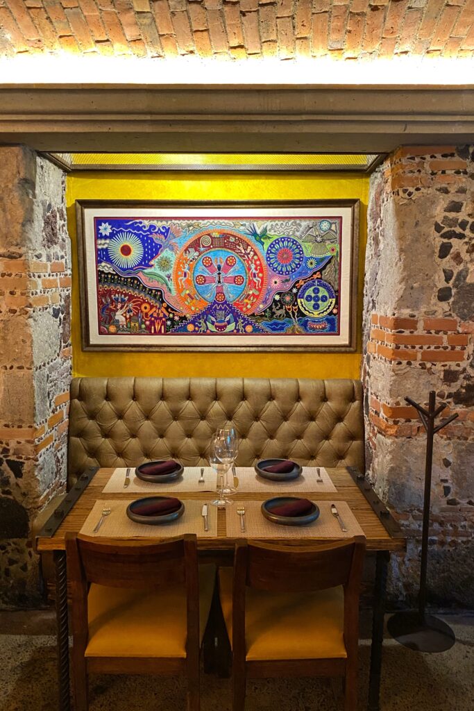 Cozy dining nook inside Los Limosneros restaurant in Mexico City’s Centro Histórico, featuring a tufted leather banquette, rustic wood table, and a vibrant Huichol-style artwork framed against a mustard-yellow wall and exposed volcanic stone. The space blends traditional Mexican craftsmanship with contemporary design — an ideal spot for travelers seeking aesthetic restaurants and cultural dining experiences in Mexico City.