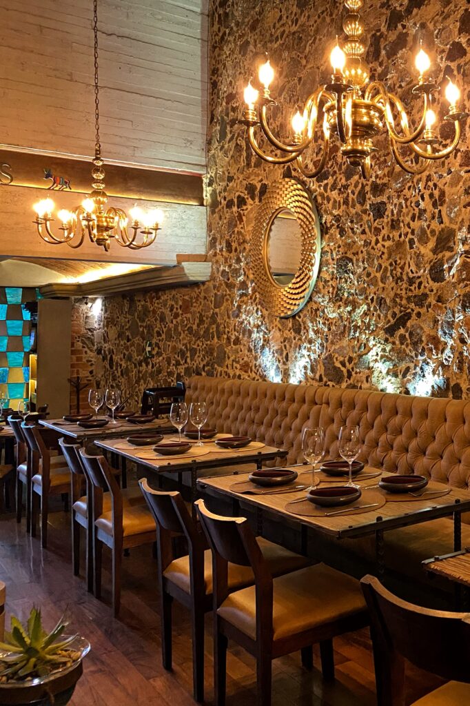 Elegant dining room at Los Limosneros in Mexico City’s Centro Histórico, featuring tufted leather banquette seating, volcanic stone walls, brass chandeliers, and rustic wooden tables set with minimalist Mexican dishware. A warm, design-rich restaurant interior that blends heritage architecture with modern sophistication — one of the most aesthetic dining spaces in Mexico City.