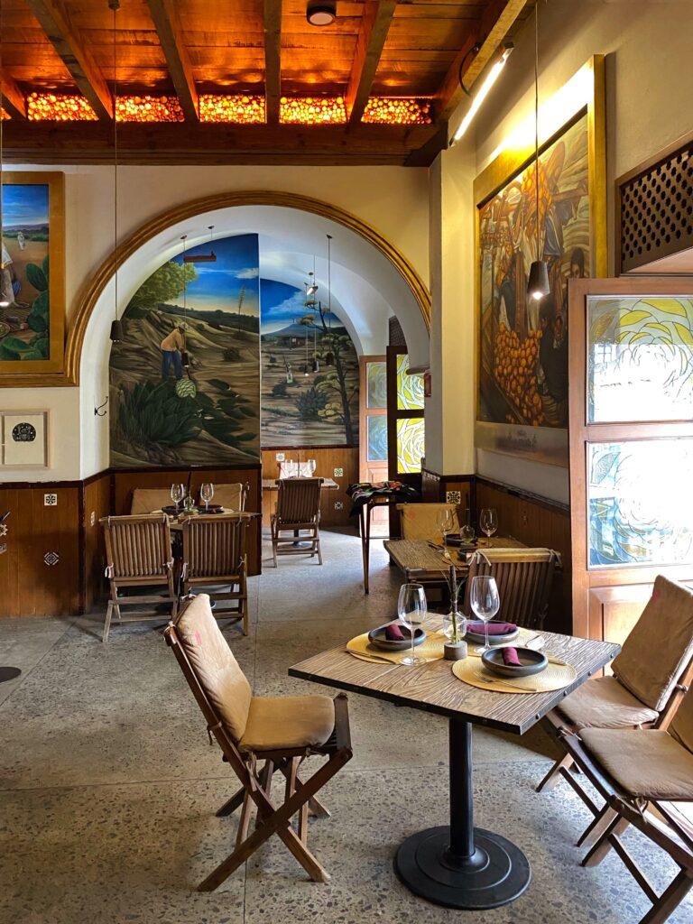 Interior dining room at Los Limosneros restaurant in Mexico City’s Centro Histórico, featuring hand-painted murals inspired by Mexican agriculture, arched doorways trimmed in gold, and a wood-beamed ceiling illuminated by warm amber light. Rustic tables are set with minimalist dishware and natural textures, creating a sophisticated yet soulful restaurant design in the heart of Mexico City.