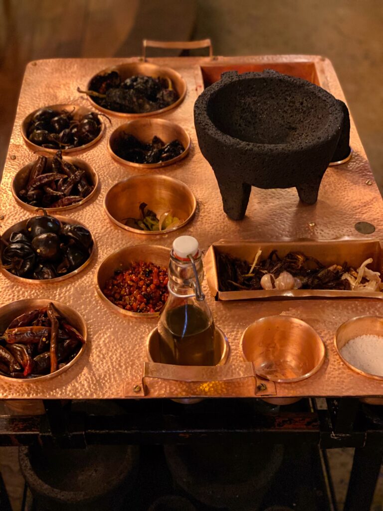 Close-up of a handcrafted copper tray filled with dried chiles, spices, and roasted ingredients beside a traditional volcanic stone molcajete at Los Limosneros restaurant in Mexico City’s Centro Histórico. This tableside salsa preparation showcases authentic Mexican culinary techniques and artisanal materials, blending design and gastronomy for an immersive dining experience in one of Mexico City’s top restaurants.