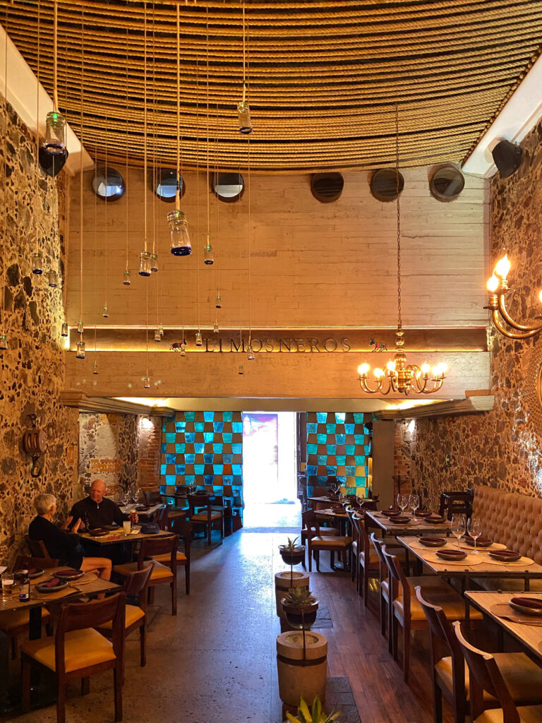Dining room interior of Los Limosneros restaurant in Mexico City’s Centro Histórico, showcasing volcanic stone walls, warm wood tables, leather seating, and a ceiling wrapped in thick woven rope with hanging glass vessels. The space combines rustic Mexican materials with modern design for a sophisticated dining experience in one of Mexico City’s most aesthetic restaurants.