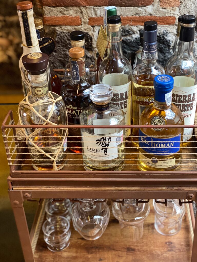 Curated spirits cart at Los Limosneros restaurant in Mexico City’s Centro Histórico, featuring premium bottles of mezcal, whisky, and artisanal Mexican liquors arranged on a rustic metal and wood trolley against a volcanic stone and brick wall. A refined design detail that reflects Mexico City’s elevated cocktail culture and craftsmanship-driven dining scene.