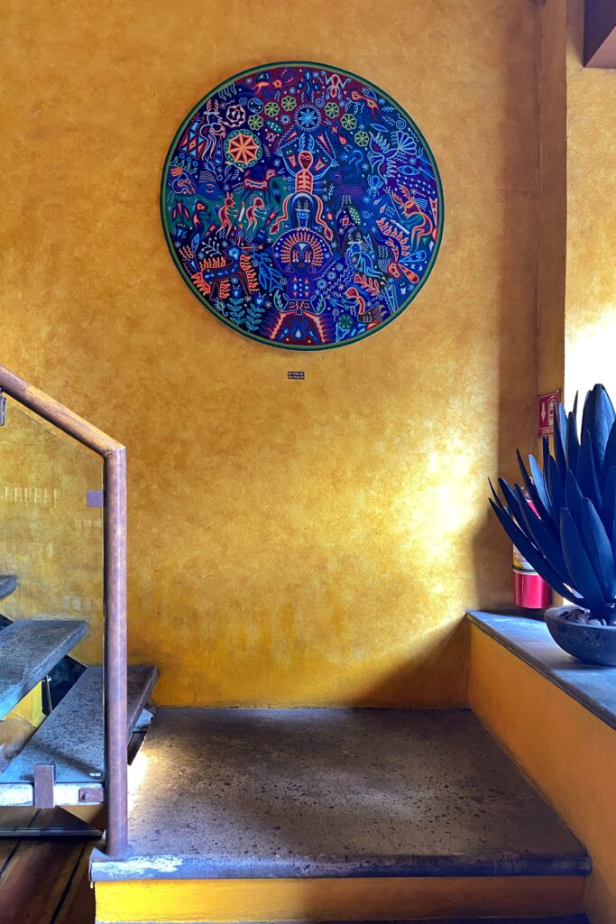Stairwell at Los Limosneros restaurant in Mexico City’s Centro Histórico, featuring a vibrant circular Huichol-style artwork mounted on a mustard-yellow wall, beside a sculptural agave plant in a stone pot. The space highlights modern Mexican design rooted in indigenous artistry and bold color — a hallmark of aesthetic travel and interior inspiration in Mexico City.