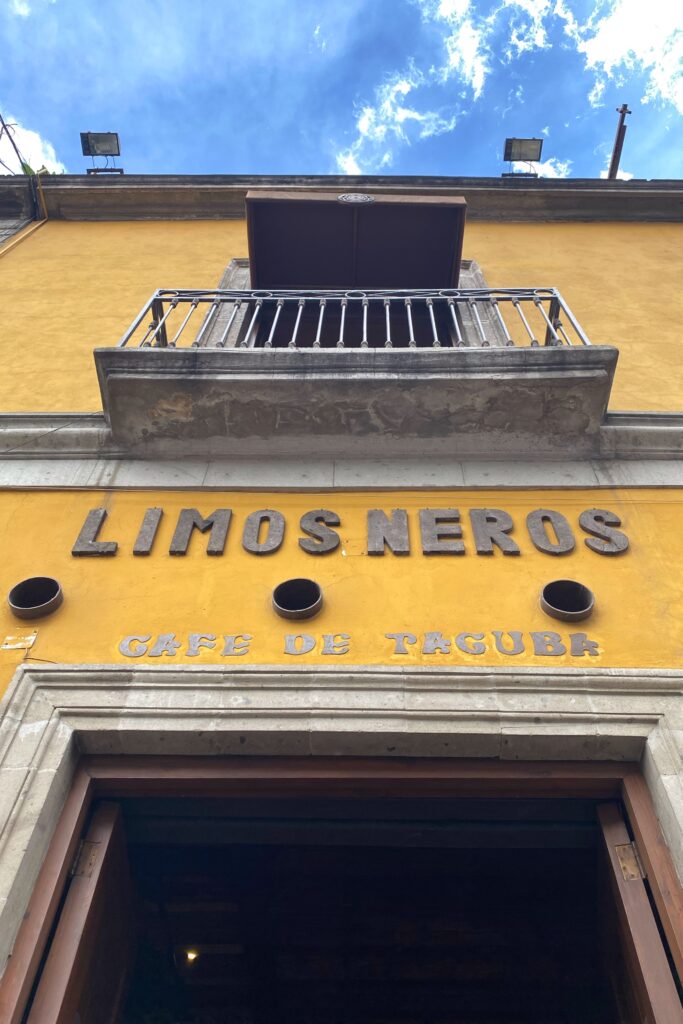 Exterior of Los Limosneros restaurant in Mexico City’s Centro Histórico, featuring a mustard-yellow colonial façade, carved stone balcony, and rustic metal signage under a bright blue sky — a mix of heritage architecture and modern Mexican design, ideal for travelers exploring Mexico City’s best restaurants and aesthetic dining spots.