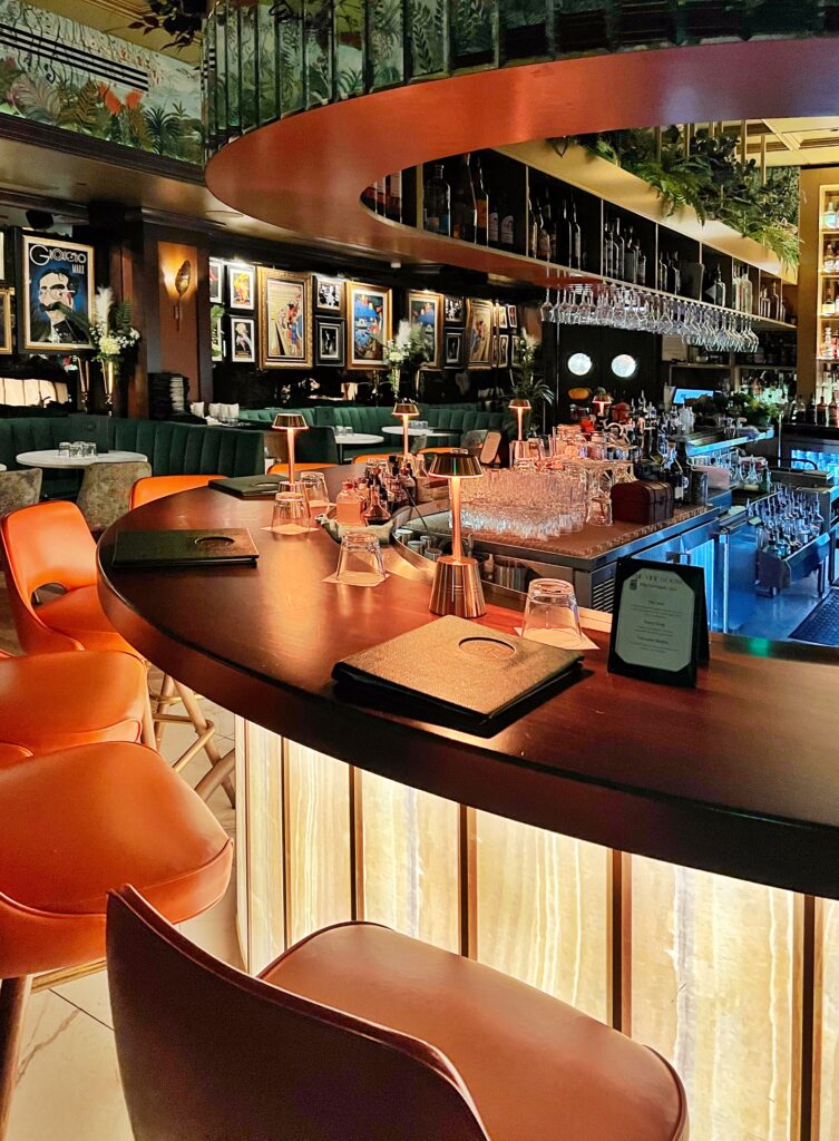 The Vine Room in Naples, Florida — a moody bar aesthetic, luxurious speakeasy bar featuring warm amber lighting, velvet green banquettes, mid-century bar stools, and curated art decor, creating an elegant and intimate cocktail lounge atmosphere.