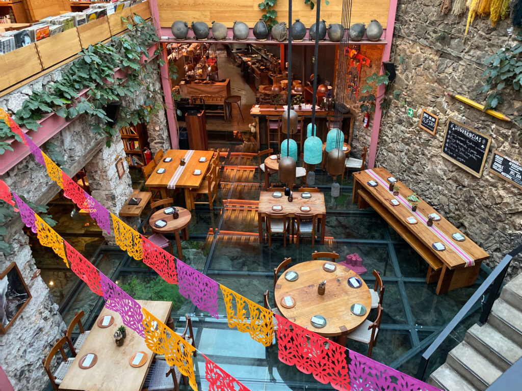 Interior view of Tetetlán café in Mexico City showing the glass-floor dining area, rustic stone walls, wood furniture, and colorful papel picado banners — a striking architectural and cultural destination featured in the Roam & Reside Mexico City Aesthetic Travel Guide.