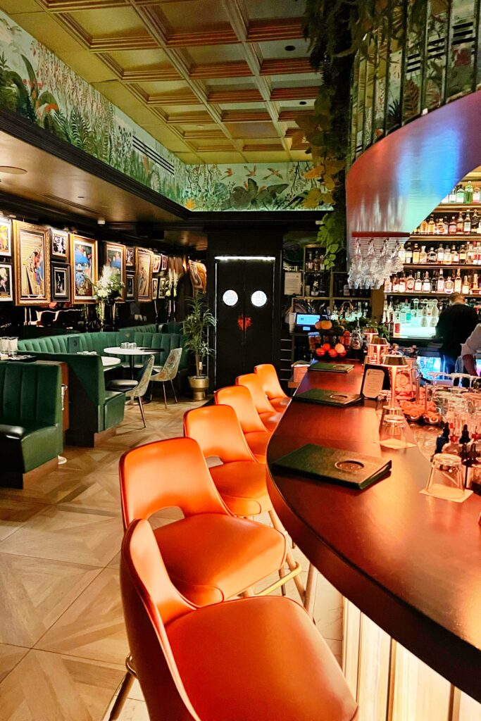Interior of The Vine Room speakeasy in Naples, Florida, featuring rich green velvet banquettes, coral barstools, tropical mural ceiling, and art-filled walls creating a modern, maximalist vintage cocktail lounge aesthetic.