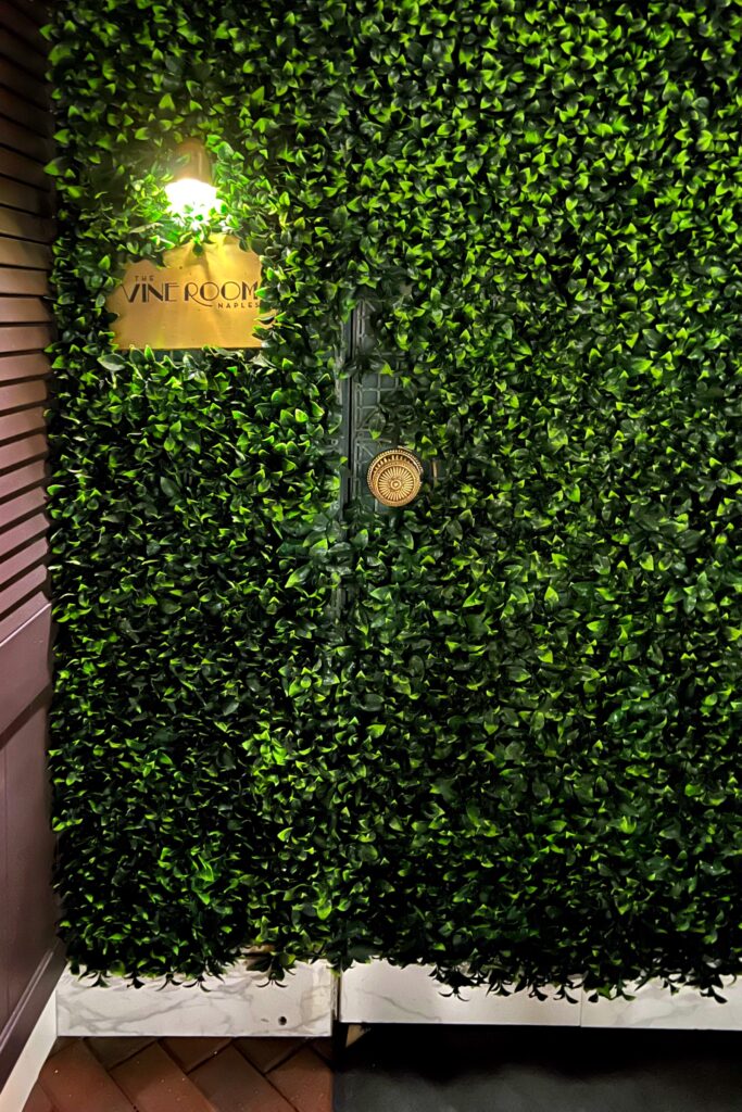 Hidden entrance to The Vine Room in Naples, Florida, featuring a lush green foliage wall with a brass plaque and gold doorknob — a stylish speakeasy bar entrance blending luxury design and mystery.