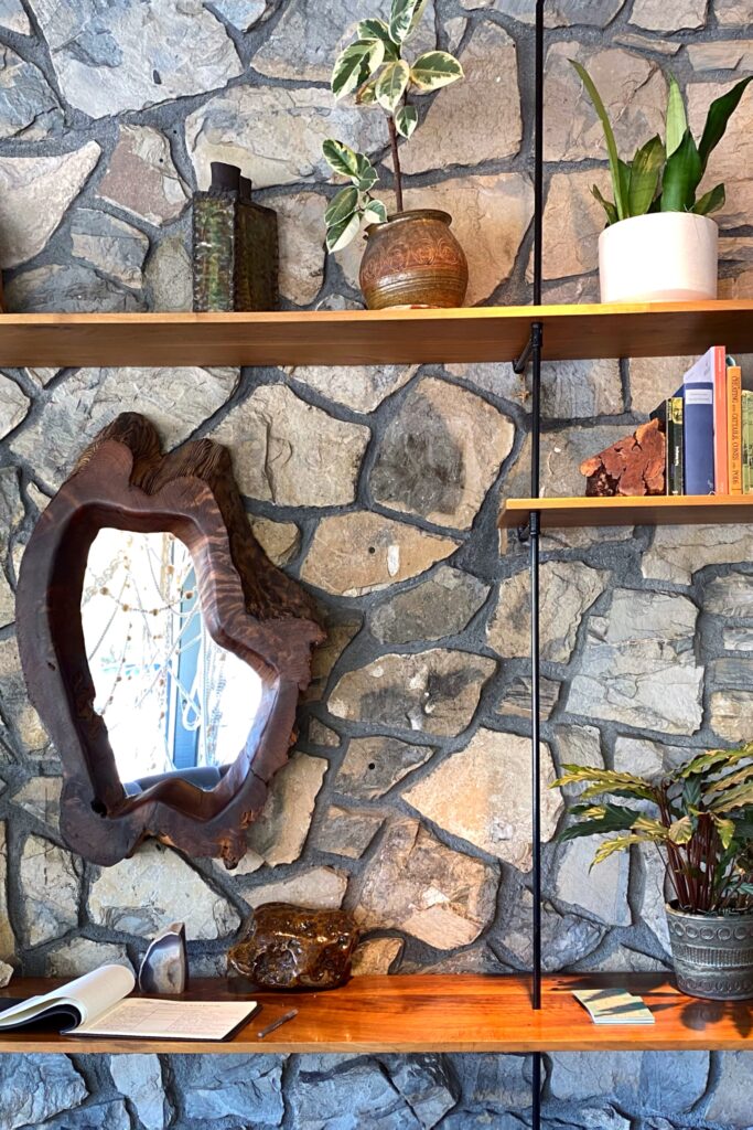 Stone-wall vignette at the Ace Hotel in Palm Springs featuring floating wood shelves styled with potted plants, ceramic vessels, vintage books, and an organic wood-framed mirror — a warm, desert-modern interior design moment.