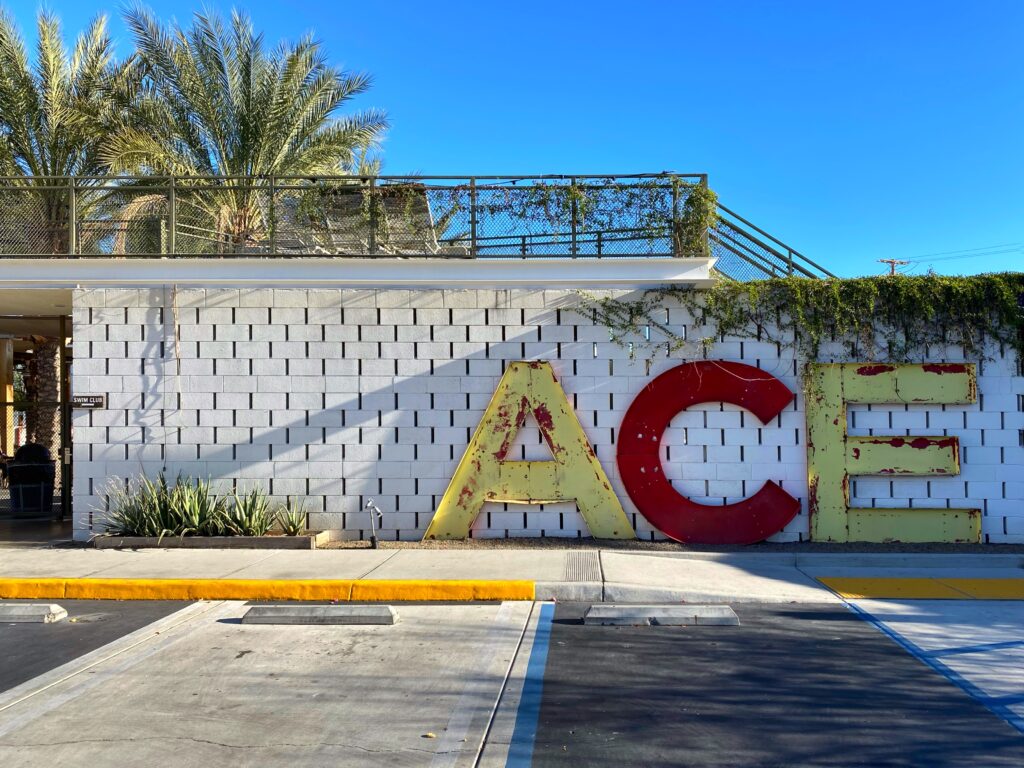 Image from the Palm Springs Aesthetic Travel Guide. Vintage ‘ACE’ sign against a white block wall at the Ace Hotel Palm Springs, framed by desert plants and clear blue sky — an iconic desert-modern entrance perfect for design-minded travelers.