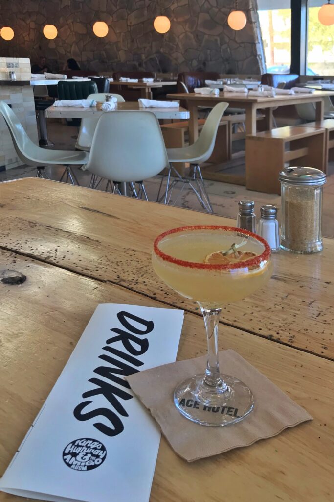 Signature cocktail at Kings Highway restaurant inside the Ace Hotel Palm Springs, served on a rustic wood table with the dining room’s midcentury chairs, stone wall, and globe pendant lights in the background.