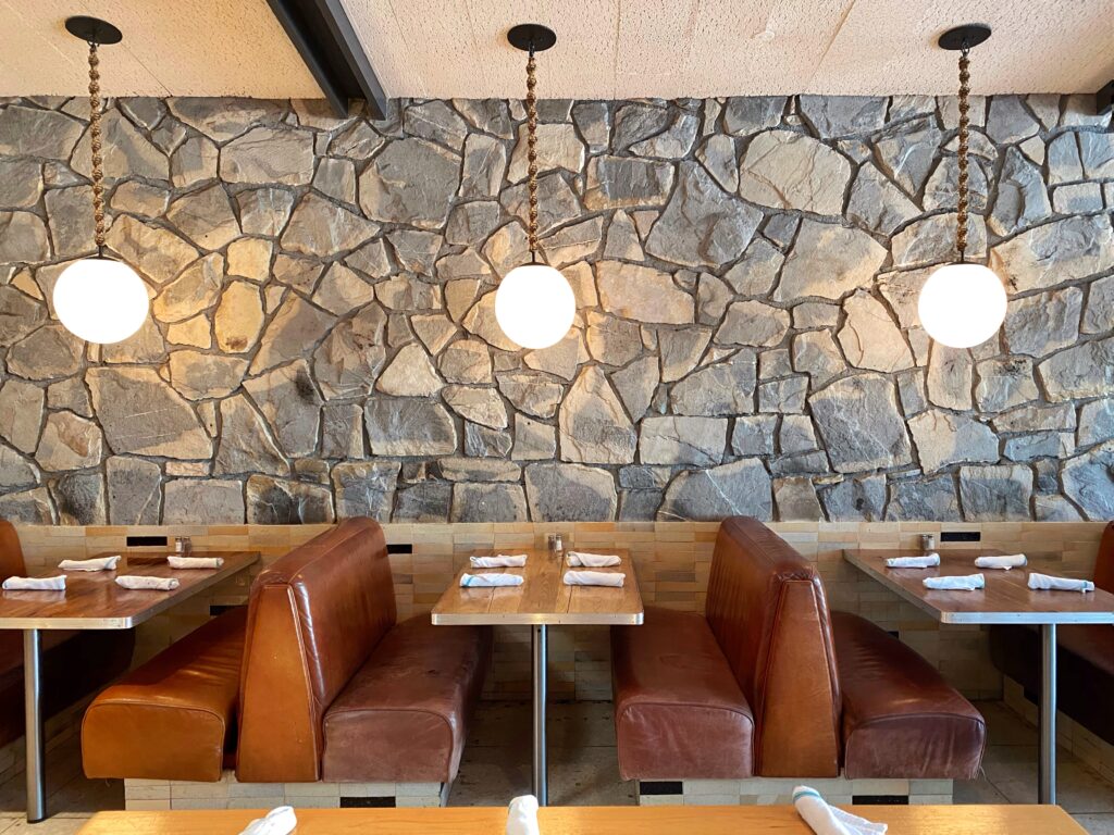 Kings Highway restaurant at the Ace Hotel Palm Springs, featuring caramel leather diner booths, a dramatic stone accent wall, and globe pendant lights — a modern take on midcentury desert-diner design. Image from the Palm Springs Aesthetic Travel Guide by Roam & Reside.
