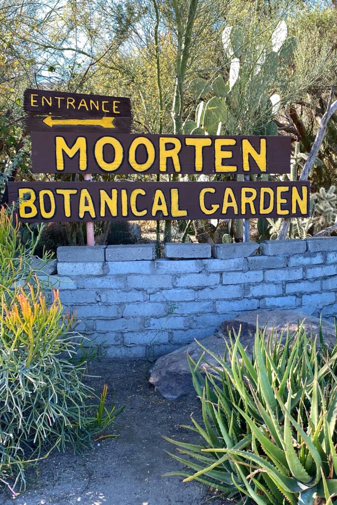 Entrance sign for Moorten Botanical Garden in Palm Springs, displayed on rustic wooden boards with yellow lettering, surrounded by desert plants, cacti, and a low brick wall.