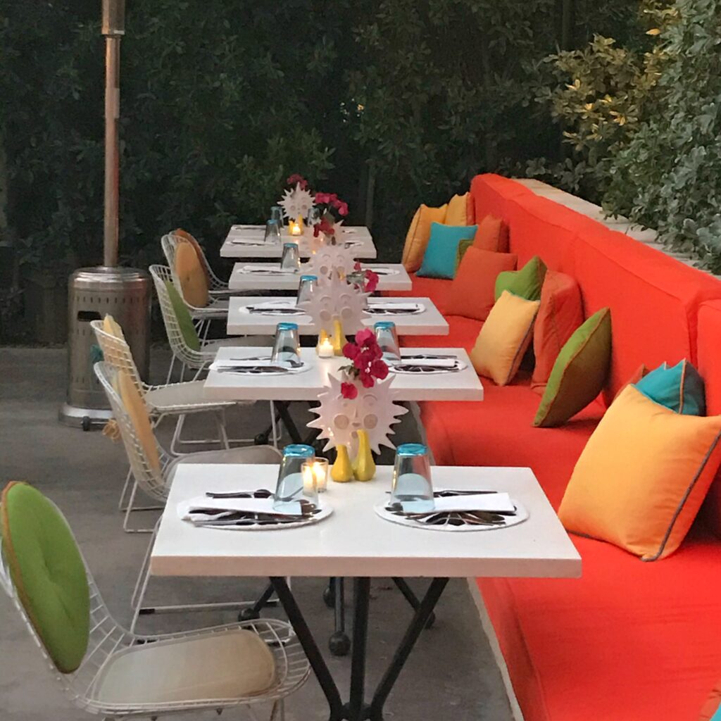 Close-up view of Norma’s at the Parker Palm Springs featuring bright orange banquette seating, colorful pillows, white patio tables, and midcentury wire chairs set for outdoor dining.