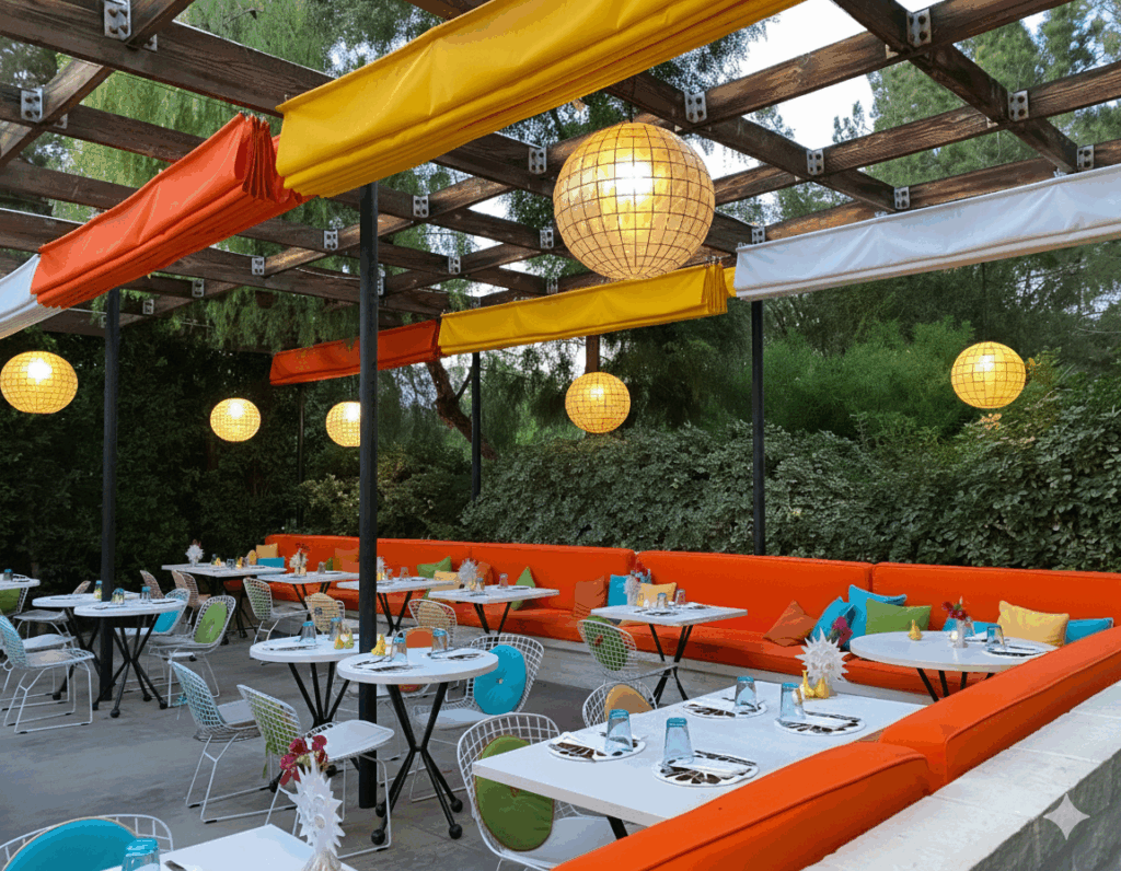 Colorful outdoor patio at Norma’s at the Parker Palm Springs featuring bright orange banquette seating, midcentury wire chairs, globe pendant lights, and a lush garden backdrop.