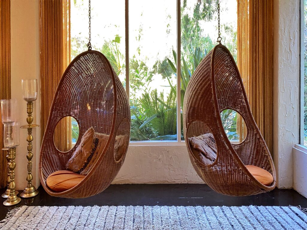 Pair of hanging rattan pod chairs with orange cushions in the lobby at the Parker Palm Springs, surrounded by brass candleholders and floor-to-ceiling windows overlooking lush greenery.