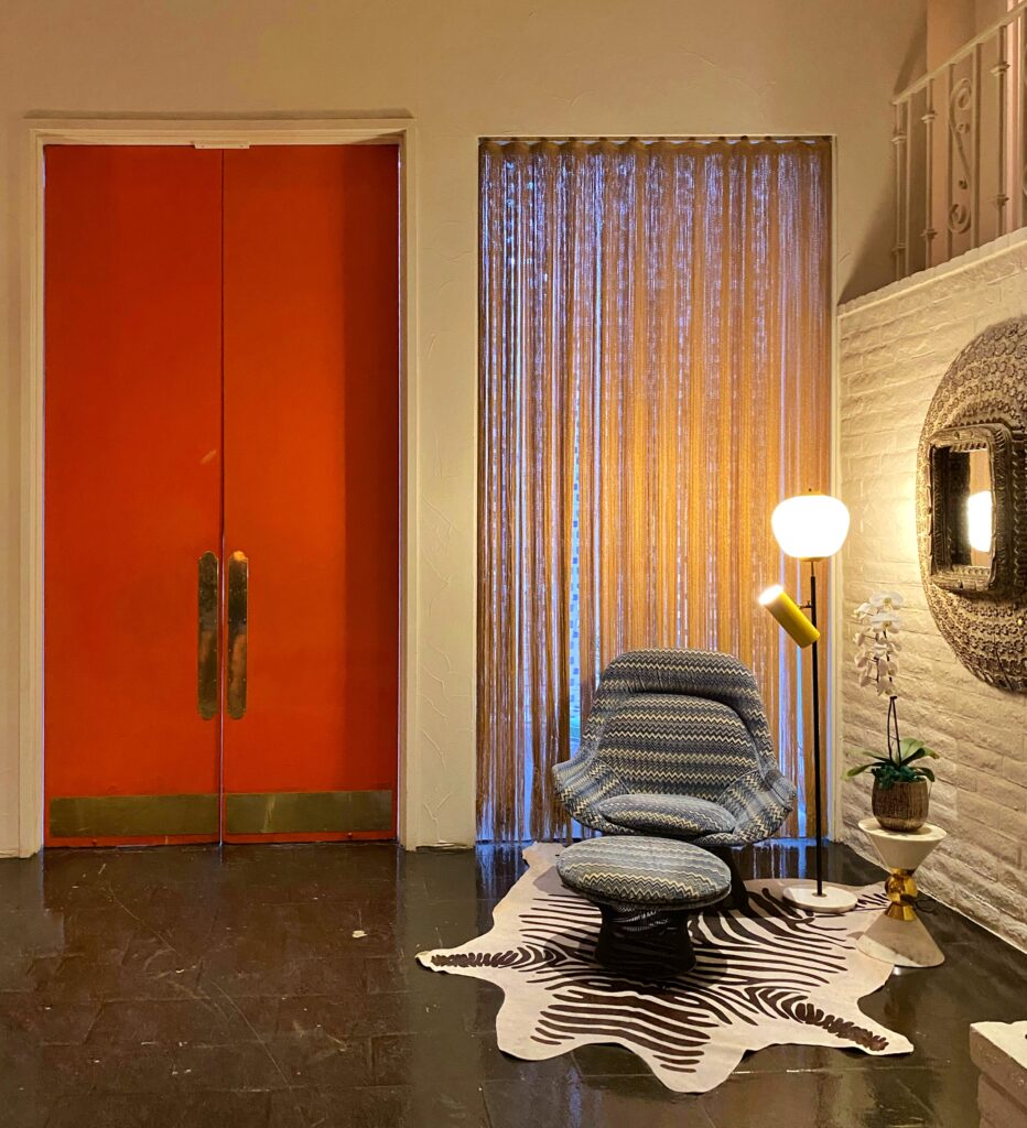 Midcentury-inspired reading nook at the Parker Palm Springs with bold orange double doors, a patterned lounge chair and ottoman on a zebra rug, gold floor lamp, and eclectic decor.