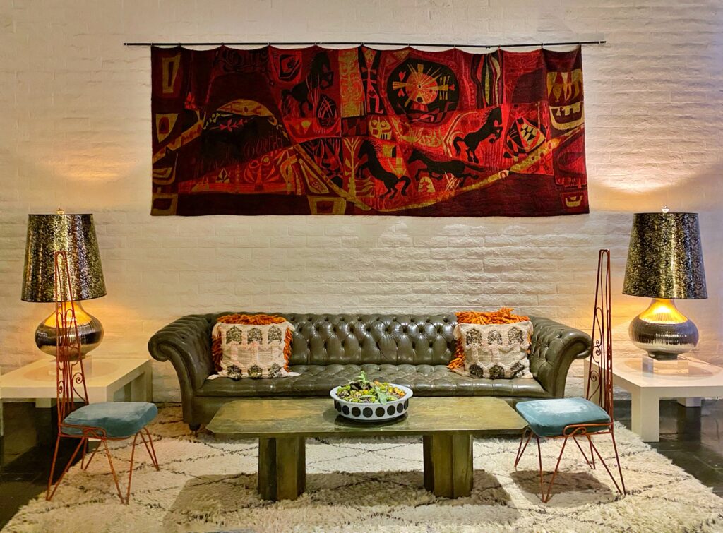 Vintage-inspired lounge at the Parker Palm Springs featuring a tufted leather sofa, midcentury lamps, colorful woven tapestry, and eclectic desert-modern décor.