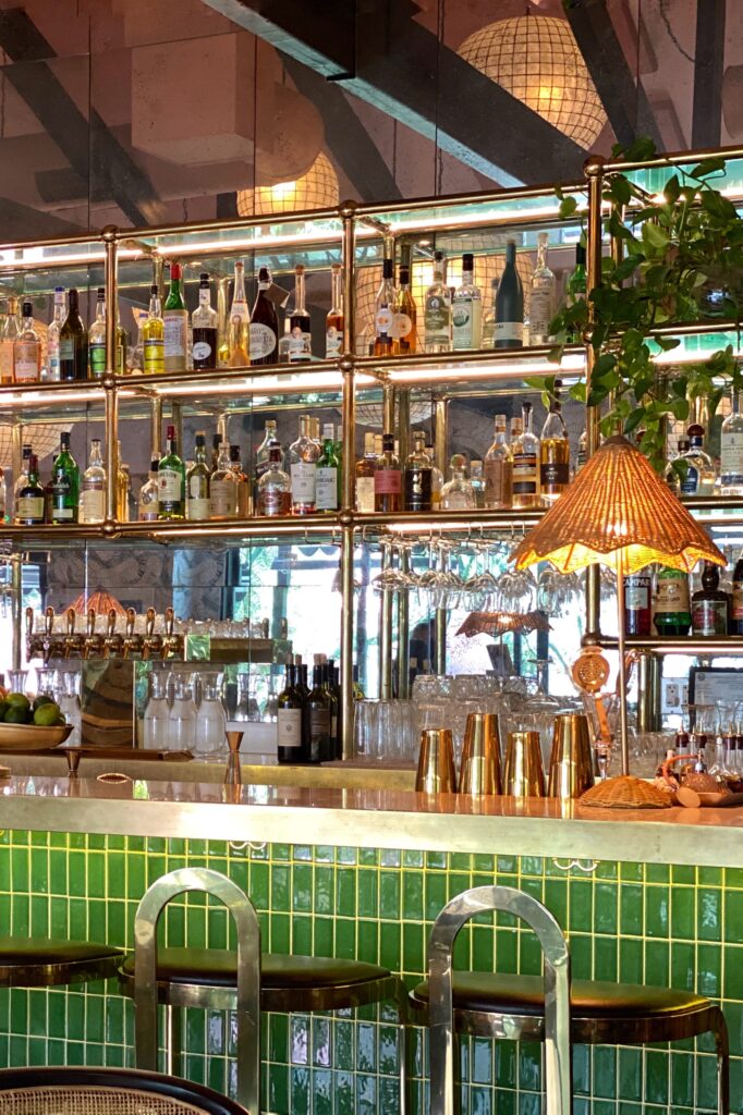 Elegant Pink Cabana bar in Palm Springs with glossy green tile, brass shelving filled with spirits, rattan lamp, hanging glassware, and mirrored back wall reflecting globe lights.