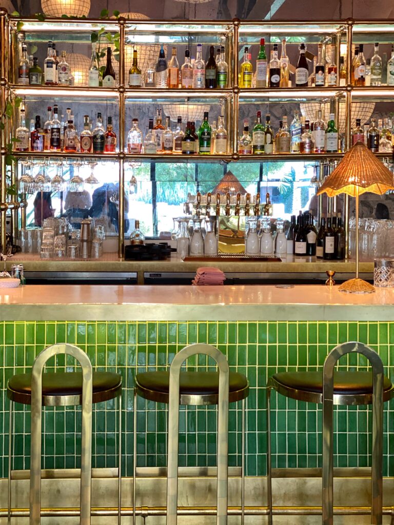 Front view of the Pink Cabana bar in Palm Springs featuring glossy green tile, brass shelving lined with spirits, rattan lamp, hanging glassware, and modern metal bar stools.
