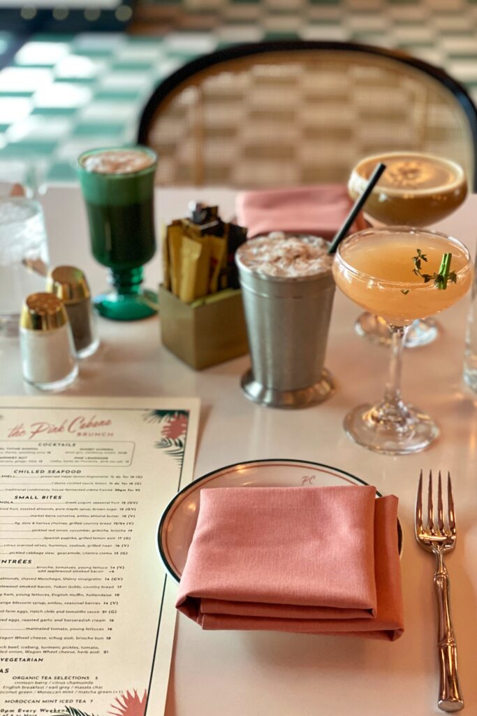 Brunch table at the Pink Cabana in Palm Springs with pastel napkins, bamboo-style flatware, tropical cocktails, iced drinks, and the brunch menu on a white table against the restaurant’s green-and-white tiled floor.
