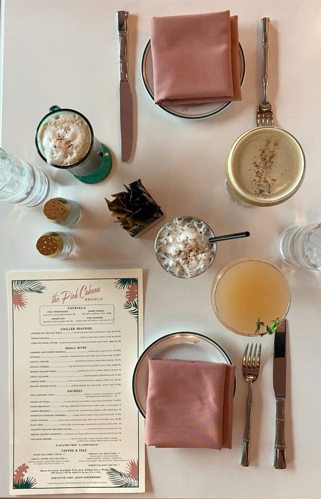 Brunch flatlay at the Pink Cabana restaurant in Palm Springs, featuring pastel napkins, bamboo-style cutlery, specialty cocktails, iced drinks, and the printed brunch menu on a modern white table.